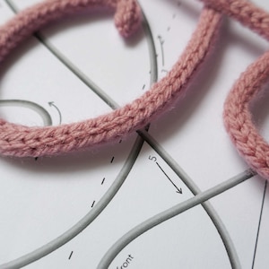 May include: Close-up of a pink knitted cord being used to create a letter on a white paper pattern with gray lines and arrows.