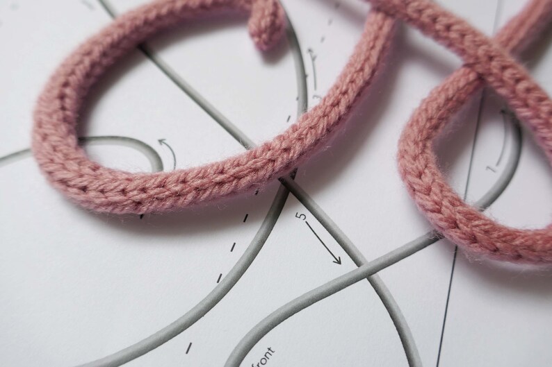 May include: Close-up of a pink knitted cord being used to create a letter on a white paper pattern with gray lines and arrows.