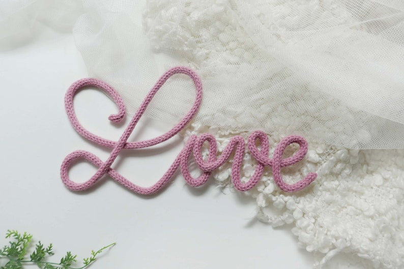 May include: Pink yarn letters spelling out the word "Love" on a white background.