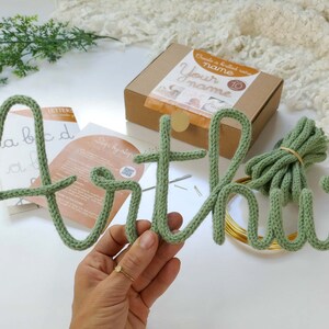 May include: A sage green knitted name, "Arthur", is shown, with the letters made from thick yarn. The name is accompanied by a kit in a brown box, with the text "Create a knitted wire Your name". Additional materials include wire and instructions.