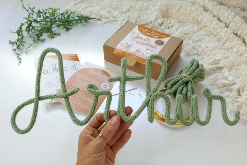 May include: A sage green knitted name, "Arthur", is shown, with the letters made from thick yarn. The name is accompanied by a kit in a brown box, with the text "Create a knitted wire Your name". Additional materials include wire and instructions.