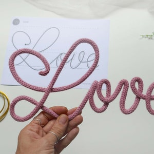 May include: A pink knitted word "Love" is displayed on a white surface. The word is made of a thick, soft yarn and is shaped in a cursive font.