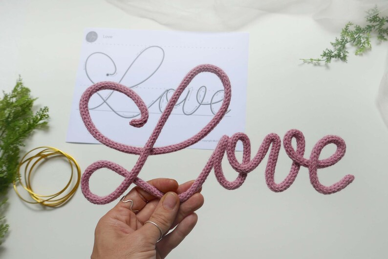May include: A pink knitted word "Love" is displayed on a white surface. The word is made of a thick, soft yarn and is shaped in a cursive font.