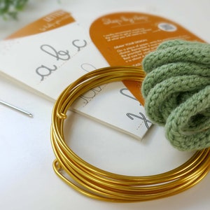 May include: A craft kit featuring gold wire, green yarn, needles, and lettering guides. The kit includes a needle, pins, and a spool of gold wire, ideal for craft projects and DIY enthusiasts.