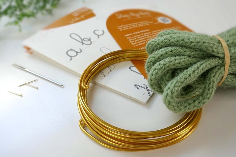 May include: A craft kit featuring gold wire, green yarn, needles, and lettering guides. The kit includes a needle, pins, and a spool of gold wire, ideal for craft projects and DIY enthusiasts.