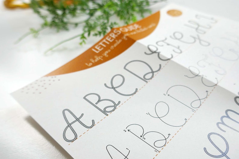 May include: A calligraphy guide showing the letters A, B, D, E, and H in a cursive style. The guide includes instructions on how to create each letter. The guide is white with grey lettering and an orange accent.