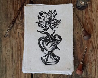 May include: A black and white woodcut print of a stylized flower with snakes wrapped around a chalice. The print is on a sheet of paper and is surrounded by a wooden surface.