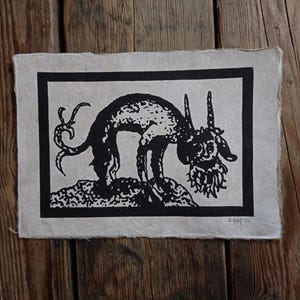 Pode incluir: Impressão em preto e branco de uma criatura com chifres e barba, em pé sobre uma superfície texturizada. A obra de arte é emoldurada com uma borda preta e está em papel de cor clara. A impressão é assinada e numerada.