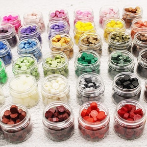 May include: Assortment of 30 small glass jars filled with colorful wax beads. The jars are arranged in rows on a white surface. The wax beads are in various colors, including red, green, blue, yellow, and white.