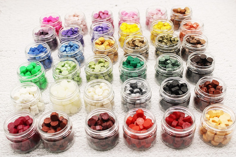 May include: Assortment of 30 small glass jars filled with colorful wax beads. The jars are arranged in rows on a white surface. The wax beads are in various colors, including red, green, blue, yellow, and white.