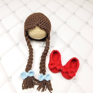 May include: A crochet doll head with brown braids and light blue bows, paired with two red crochet baby shoes. The doll head has a tan face and brown hair. The shoes have a bow on the front.