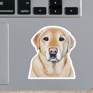 May include: A sticker featuring a detailed illustration of a yellow Labrador Retriever dog. The dog has brown eyes and a pink nose. The sticker is placed on a laptop, with a wooden surface in the background.
