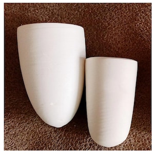 May include: Two unglazed ceramic vessels with a matte off-white finish. The larger vessel features a tapered, rounded base, while the smaller vessel has a more cylindrical shape with a rounded bottom. Both items have smooth, plain surfaces.