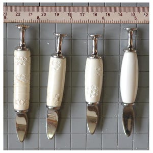 May include: Four butter knives with ivory-coloured handles and stainless steel blades. The handles have decorative patterns, and the knives are arranged against a grey grid background. The ruler shows measurements in centimetres.