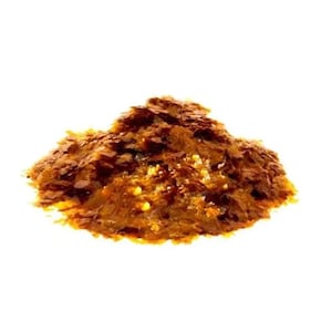 May include: A pile of small, thin, irregular flakes in shades of amber, copper, and dark brown. The material has a translucent, layered appearance with a glossy, reflective surface texture, forming a mound against a plain white background.