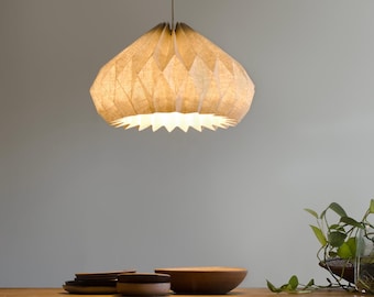 May include: A beige, origami-style pendant light fixture hangs from the ceiling. The light is on and casts a warm glow. The light fixture is made of fabric and has a pleated design.