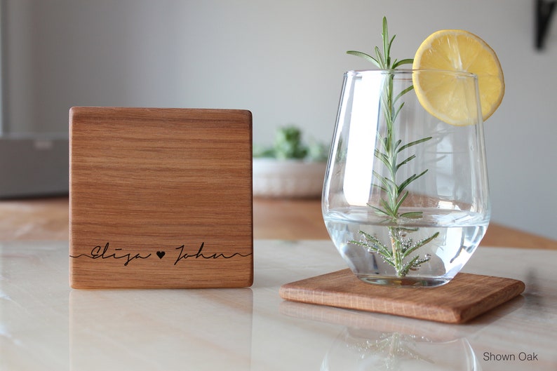 Op de afbeelding: Twee vierkante houten onderzetters met een natuurlijke houten afwerking. De onderzetter links heeft de namen "Eliza" en "John" gegraveerd met een hartje tussen de namen. De onderzetter rechts is leeg. De onderzetters worden getoond op een wit oppervlak met een glas water en een citroenschijf op de onderzetter rechts. De tekst "Shown Oak" is zichtbaar in de rechterbenedenhoek van de afbeelding.