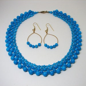 May include: A turquoise beaded necklace and matching earrings set. The necklace features multiple strands of graduated blue beads, with a gold-tone clasp. The earrings have gold-tone hoops with small turquoise beads. The jewelry set is displayed on a white surface.