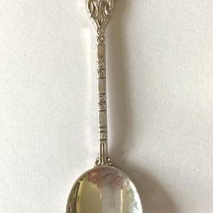 May include: A silver souvenir spoon with a detailed handle and a bowl-shaped scoop. The spoon's handle features an emblem with the word "Cornwall" and a coat of arms. The spoon is likely a collectible item.