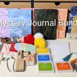 May include: A selection of journals, stationery, and craft supplies. The image shows journals with diverse covers, colourful ink pads, decorative tape, stamps, bookmarks, and other crafting accessories. The text "Mystery Journal Bundle" is displayed.