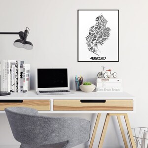 May include: A white desk with a black lamp, a laptop, a stack of books, a small potted plant, a toy bicycle, and a framed black and white print of the word "JERSEY CITY" in a stylized font that resembles the shape of the city.