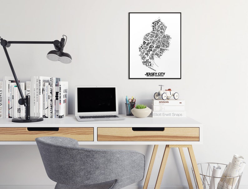 May include: A white desk with a black lamp, a laptop, a stack of books, a small potted plant, a toy bicycle, and a framed black and white print of the word "JERSEY CITY" in a stylized font that resembles the shape of the city.