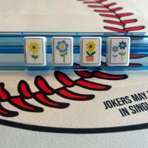 May include: Four white rectangular tiles with floral designs, each featuring a different flower in yellow and blue, are displayed in a blue plastic holder. The background includes a baseball-themed design with red stitching and the text "JOKERS MAY IN SING".