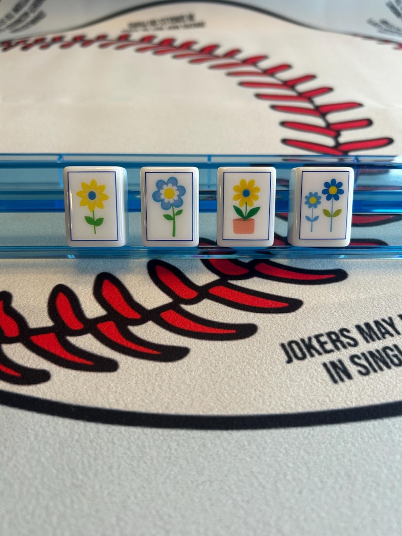 May include: Four white rectangular tiles with floral designs, each featuring a different flower in yellow and blue, are displayed in a blue plastic holder. The background includes a baseball-themed design with red stitching and the text "JOKERS MAY IN SING".