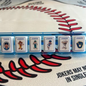 May include: A close-up shot of a baseball-themed game. Six white tiles with the word "JOKER" and cartoon baseball player illustrations are in a blue tray. The background features a baseball design with red stitching.