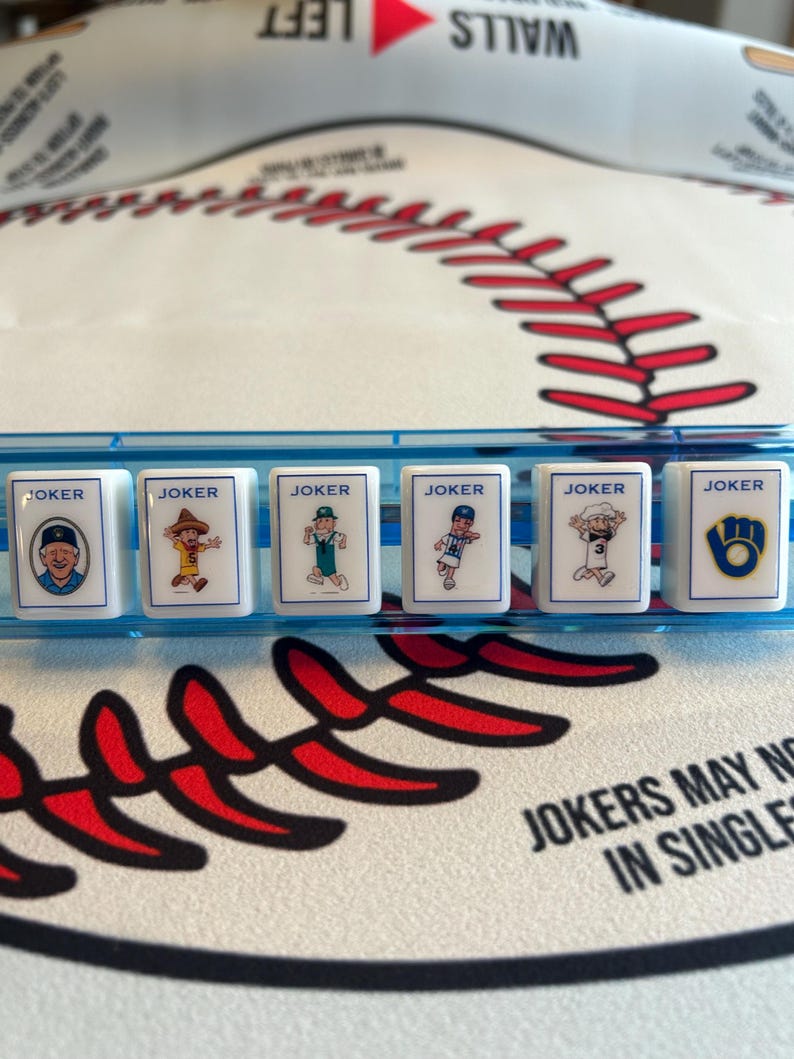 May include: A close-up shot of a baseball-themed game. Six white tiles with the word "JOKER" and cartoon baseball player illustrations are in a blue tray. The background features a baseball design with red stitching.