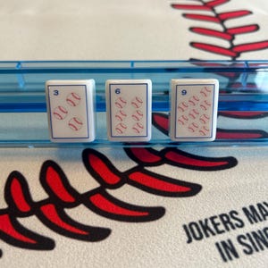 May include: Three white game cards with baseball graphics, each displaying a number and baseballs. The cards are in a blue holder, set against a baseball-themed background with red stitching and the text "JOKERS MAY IN SING".