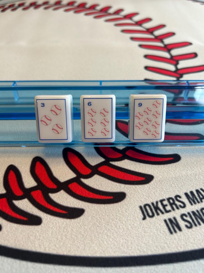 May include: Three white game cards with baseball graphics, each displaying a number and baseballs. The cards are in a blue holder, set against a baseball-themed background with red stitching and the text "JOKERS MAY IN SING".