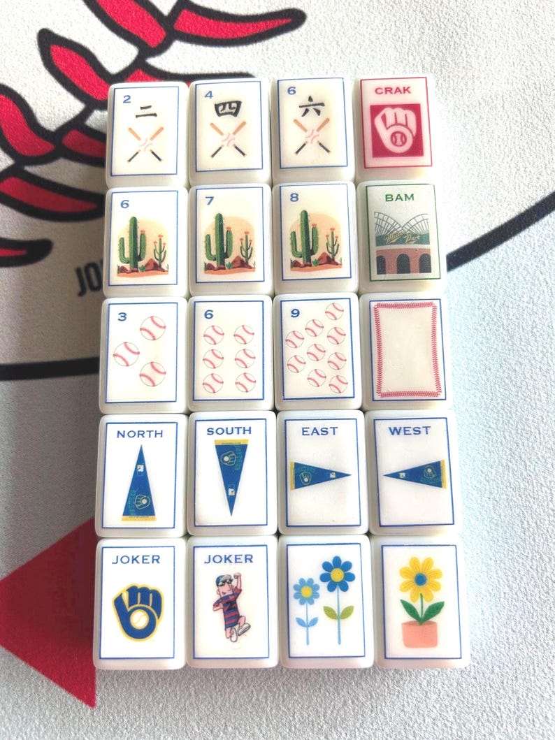 May include: A collection of white Mahjong tiles with various illustrations. The tiles feature numbers, baseballs, cacti, and directional markers. Some tiles have the words "CRAK", "BAM", "NORTH", "SOUTH", "EAST", "WEST", and "JOKER".