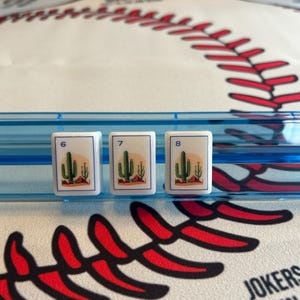 May include: Three white game tiles with the numbers 6, 7, and 8. Each tile features a desert scene with a cactus and a setting sun. The tiles are set against a blue background, with a baseball-themed surface.