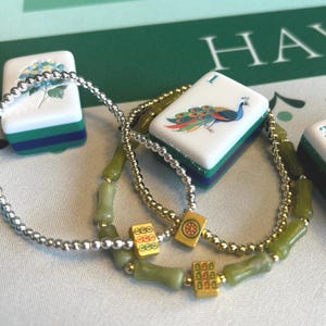 May include: Close-up of two beaded necklaces with green and gold accents, and several Mahjong tiles. The tiles are white with colorful designs, including a peacock and a blue flower. The background is a green surface.