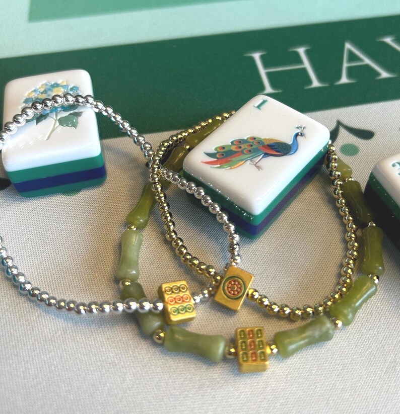 May include: Close-up of two beaded necklaces with green and gold accents, and several Mahjong tiles. The tiles are white with colorful designs, including a peacock and a blue flower. The background is a green surface.