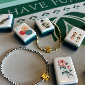 May include: A collection of white Mahjong tiles with colorful illustrations, including a peacock, umbrellas, and flowers. Two beaded bracelets with gold and silver beads are also present. The background features the text "HAVE FUN" on a green surface.