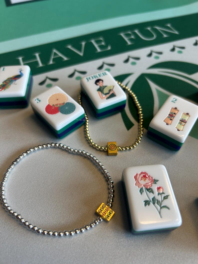 May include: A collection of white Mahjong tiles with colorful illustrations, including a peacock, umbrellas, and flowers. Two beaded bracelets with gold and silver beads are also present. The background features the text "HAVE FUN" on a green surface.