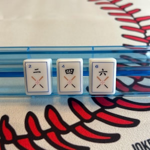 May include: Three white Mahjong tiles with black symbols for the numbers 2, 4, and 6. The tiles are in a blue plastic holder, set against a baseball-themed background with red stitching.