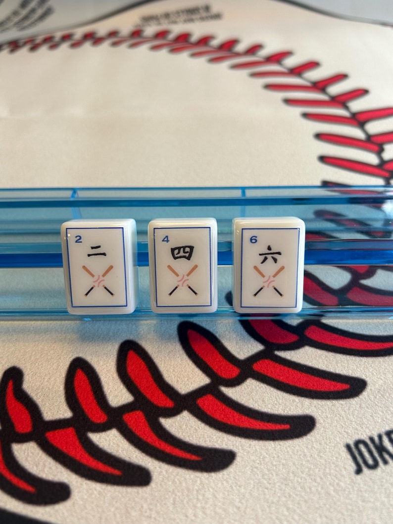 May include: Three white Mahjong tiles with black symbols for the numbers 2, 4, and 6. The tiles are in a blue plastic holder, set against a baseball-themed background with red stitching.