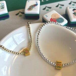 May include: Two beaded bracelets, one gold and one silver, rest on a white plate. Each bracelet features a yellow charm with a red and green design. Mahjong tiles with colorful illustrations are in the background.