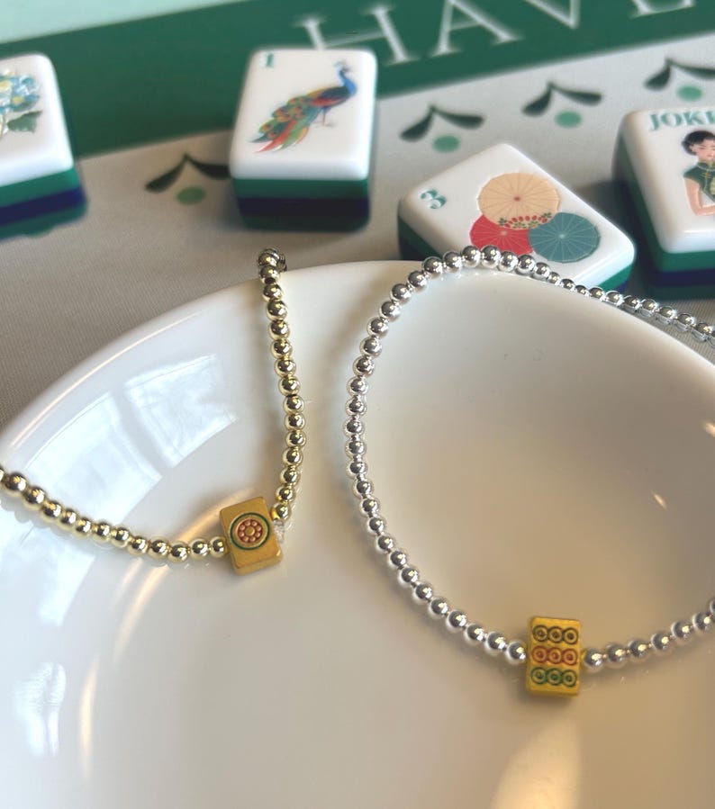 May include: Two beaded bracelets, one gold and one silver, rest on a white plate. Each bracelet features a yellow charm with a red and green design. Mahjong tiles with colorful illustrations are in the background.