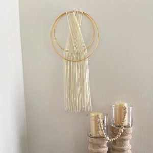 May include: A white macrame wall hanging with a gold metal hoop design. The hanging is made with a thick white yarn and has a fringe at the bottom. The hanging is displayed on a white wall with two glass candle holders with wooden bases and white candles.