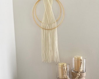 May include: A white macrame wall hanging with a gold metal hoop design. The hanging is made with a thick white yarn and has a fringe at the bottom. The hanging is displayed on a white wall with two glass candle holders with wooden bases and white candles.