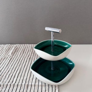 May include: Two-tiered serving stand with a silver handle. The stand is made of white ceramic with a green glaze. The stand is on a white surface with a striped tablecloth.