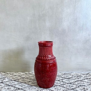 Puede incluir: Un jarrón de cerámica rojo oscuro con intrincados detalles en relieve y una superficie texturizada. El jarrón tiene una base ancha y un cuello estrecho.