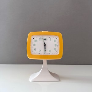 Könnte beinhalten: Ein gelb-weißer Vintage-Wecker. Die Uhr hat ein quadratisches Zifferblatt mit schwarzen Ziffern und Zeigern, und das Wort "Teter" ist auf dem Zifferblatt aufgedruckt. Die Uhr ist auf einem weißen Sockel montiert.