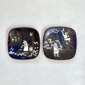 Peut inclure: Deux assiettes carrées en céramique ornées d'illustrations bleu foncé et marron. L'assiette de gauche montre une personne cuisinant avec des ours et l'année 1986. Celle de droite représente une personne atteignant un croissant de lune en 1987.