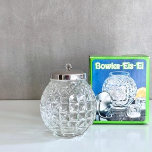 May include: A clear, faceted glass ice bowl with a silver lid and handle. The bowl is round with a diamond pattern. A box with the text "Bowlen-Eis-Ei" is behind the bowl, showing a similar bowl with ice.