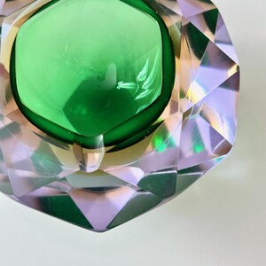 Op de afbeelding: Een decoratieve asbak in smaragdgroen en helder kristal. De asbak heeft een centrale put van groen glas, omlijst door een goudkleurige rand. De buitenkant is gesneden met geometrische facetten, waardoor het een diamantachtig uiterlijk krijgt.